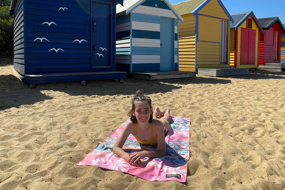 Girl lying on a pink Tesalate kids beach towel in front of colourful beach huts