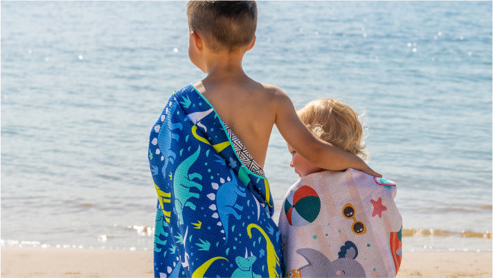 Two kids at the beach wrapped in Tesalate dinosaur and beach-print towels